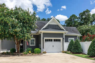 Single-story gray house with white trim, a gable roof, attached garage, and landscaped yard under a blue sky with scattered clouds.