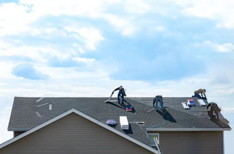 Four workers are installing or repairing shingles on the roof of a house under a partly cloudy sky.